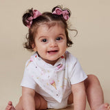 A smiling baby girl with her hair in two buns and pink bows, wearing a white bib with a pink and yellow ice cream cone pattern over a white short-sleeve bodysuit, set against a solid beige background.
