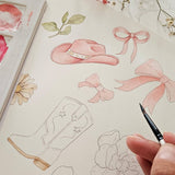 A close-up of a hand holding a paintbrush over a watercolor sketchbook featuring pink cowboy hat, bows, floral motifs, and boot outlines.