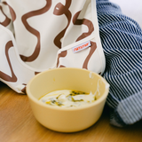 Yellow bowl of soup on a wooden table with a patterned bag and striped fabric in the background.