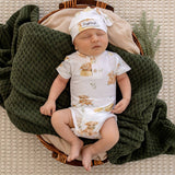 A sleeping newborn baby wearing a white short-sleeve bodysuit with teddy bear prints and a matching beanie hat, lying in a wicker basket lined with beige fabric and wrapped in a dark green textured blanket.
