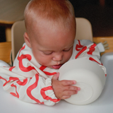 Baby in a high chair wearing a white outfit with red patterns, holding a white ball.