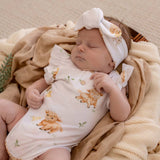 A sleeping newborn baby wearing a white short-sleeve bodysuit and matching headband, both printed with cute watercolor bison motifs, resting on a soft beige blanket.