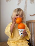 Child holding two oranges, one in each hand, with a cup in front of them.