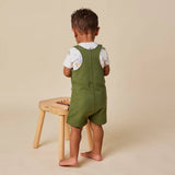 A toddler standing with their back to the camera, wearing olive green overall shorts over a white short-sleeve t-shirt with a light print. They are standing barefoot next to a small wooden stool against a solid beige background.