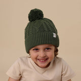 Child wearing a green knit beanie with a pom-pom on a beige background