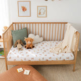 A wooden baby crib with a white mattress covered in a small animal print sheet, styled with a green pillow, plush toys, a cream cloud pillow, and a beige throw blanket in a bright nursery setting.