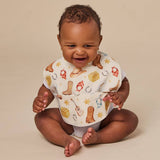 A smiling baby sitting cross-legged against a warm beige studio background, wearing a white bib printed with cowboy boots, horseshoes, stars, and small western-themed illustrations.