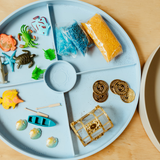 Blue tray with various toys and items including fish, a boat, and gold coins on a wooden surface.