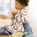 Child wearing a patterned outfit holding a cup with a bowl and spoon on a table.
