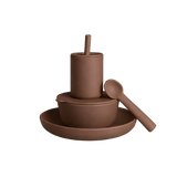 A minimalist ceramic dinnerware set in a matte terracotta brown color, featuring a deep plate, a bowl with a lid, a tall cup with a straw, and a spoon, all arranged on a solid white background.