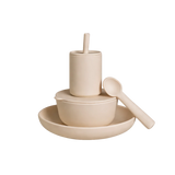 A minimalist ceramic dinnerware set in a warm cream or light beige tone, featuring a shallow plate, a bowl with a matching lid, a small cylindrical cup with a lid and straw, and a spoon resting on the side, all photographed on a seamless white background.