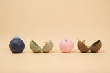 Four small capsule-shaped containers arranged in a row on a warm beige background. Each capsule is split open to reveal a small white item inside, including a dark blue one, a sage green one, a light pink one, and a taupe-brown one.