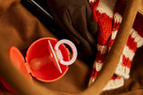 A close-up of a baby pacifier in an orange-red plastic case with a white ring handle, nestled inside a brown bag next to a red and white striped knit blanket.