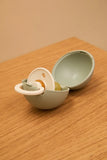 A pacifier and its matching half-shell storage case, both in a muted sage green and white color, displayed on a light wood tabletop.