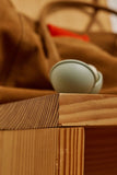 A close-up shot of a light sage green ceramic cup resting on the corner of a wooden shelf, with a brown fabric bag blurred in the background.