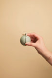 A hand holding a small, matte sage green spherical object with a small loop on top, set against a solid beige background.