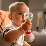 A baby holding a colorful sensory toy rattle with a clear ball and fabric ball handle, photographed in a softly lit indoor setting.