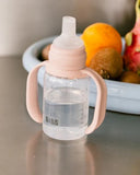 BIBS baby bottle with pink handle on a reflective surface with fruits in the background