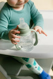 Child holding a clear sippy cup with a green handle and lid on a high chair.